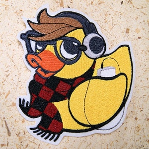May include: Embroidered iron-on patch of a yellow duck wearing a red and black checkered scarf, black framed glasses, and headphones. The duck is holding a white cup.