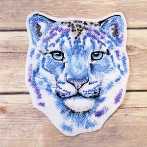 May include: Embroidered patch of a snow leopard's face. The patch is white with blue and purple embroidery. The snow leopard has blue eyes and a white nose.