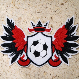 May include: Embroidered patch featuring a soccer ball within a shield, flanked by red and black wings. A crown tops the shield. The patch has a white background with red, black, and white details.