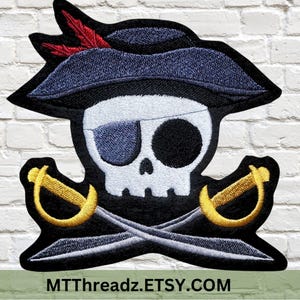 May include: Embroidered iron-on patch featuring a cartoon pirate skull wearing a black hat with a red feather, an eyepatch, and crossed swords.