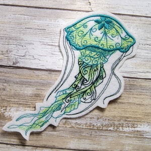 Peut inclure: Une méduse brodée en vert et bleu avec des détails complexes. La méduse a une tête en forme de cloche avec un motif floral et de longs tentacules flottants.