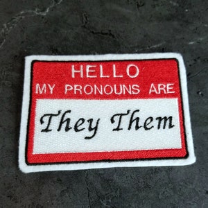 May include: Red and white name tag with the text "HELLO MY PRONOUNS ARE They Them" in black embroidery.