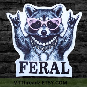 May include: A die-cut sticker featuring a black and white illustration of a raccoon wearing pink cat-eye sunglasses and layered pearl necklaces. The raccoon has both paws raised in a rock-and-roll hand gesture. The word FERAL is printed in black below.
