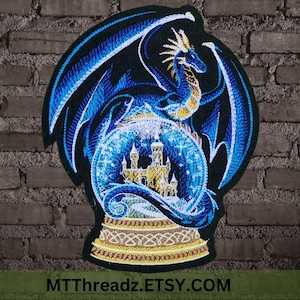 May include: Embroidered patch featuring a blue dragon curled around a snow globe with a gold castle inside. The dragon has a gold crest and white accents. The snow globe is surrounded by a gold and white border. The patch is on a black background.