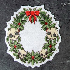 May include: A white felt Christmas wreath with green holly leaves, red berries, and four white skulls. A red bow is at the top of the wreath.