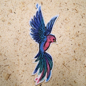 May include: Embroidered patch featuring a stylized bird with outstretched wings. The bird has a pink breast, blue wings, and a long, flowing tail with teal and purple accents. The patch is outlined in white.