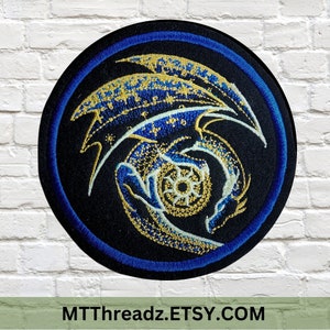 May include: A round embroidered patch featuring a blue and gold dragon design. The dragon is curled around a circle with a starburst pattern. The patch has a blue border.