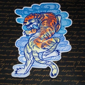May include: An embroidered iron-on patch featuring a yellow and orange tiger leaping through blue clouds. The tiger is outlined in blue and has a detailed design.