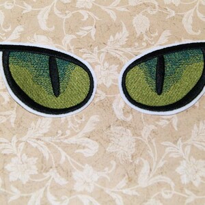K&ouml;nnte beinhalten: Zwei gr&uuml;ne gestickte Augen mit schwarzen Pupillen und wei&szlig;en Umrandungen. Die Augen befinden sich auf einem beigefarbenen Hintergrund mit einem Blumenmuster.