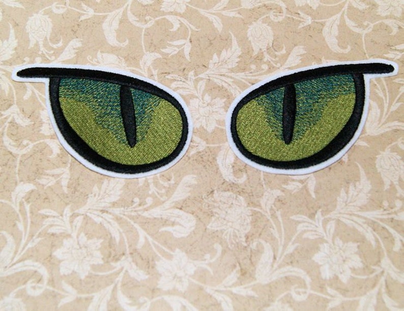 K&ouml;nnte beinhalten: Zwei gr&uuml;ne gestickte Augen mit schwarzen Pupillen und wei&szlig;en Umrandungen. Die Augen befinden sich auf einem beigefarbenen Hintergrund mit einem Blumenmuster.