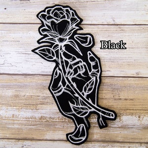 May include: Black embroidered iron-on patch featuring a hand holding a rose. The rose and hand are outlined in white thread on a black background. The word "Black" is printed in white on the right side of the patch.