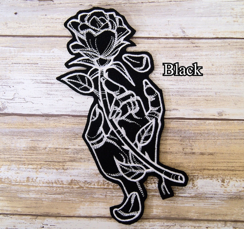 May include: Black embroidered iron-on patch featuring a hand holding a rose. The rose and hand are outlined in white thread on a black background. The word "Black" is printed in white on the right side of the patch.