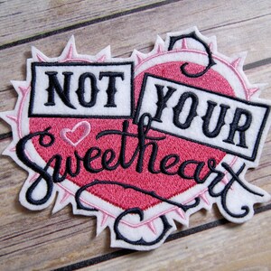 May include: A pink and white heart-shaped embroidered patch with black stitching. The patch says "Not Your Sweetheart" with a small heart in the center.