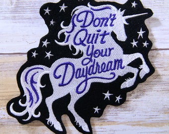 May include: White unicorn silhouette with purple embroidery on a black background. The unicorn has a horn and a flowing mane and tail. The text "Don't Quit Your Daydream" is embroidered in purple on the unicorn's body.