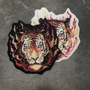May include: Embroidered patch featuring two tiger faces with flames behind them. The tigers have orange and black fur with green eyes. The flames are red and orange.