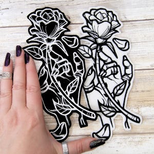 May include: Two black and white embroidered iron-on patches featuring a hand holding a rose. The rose on the left patch is black and the rose on the right patch is white.