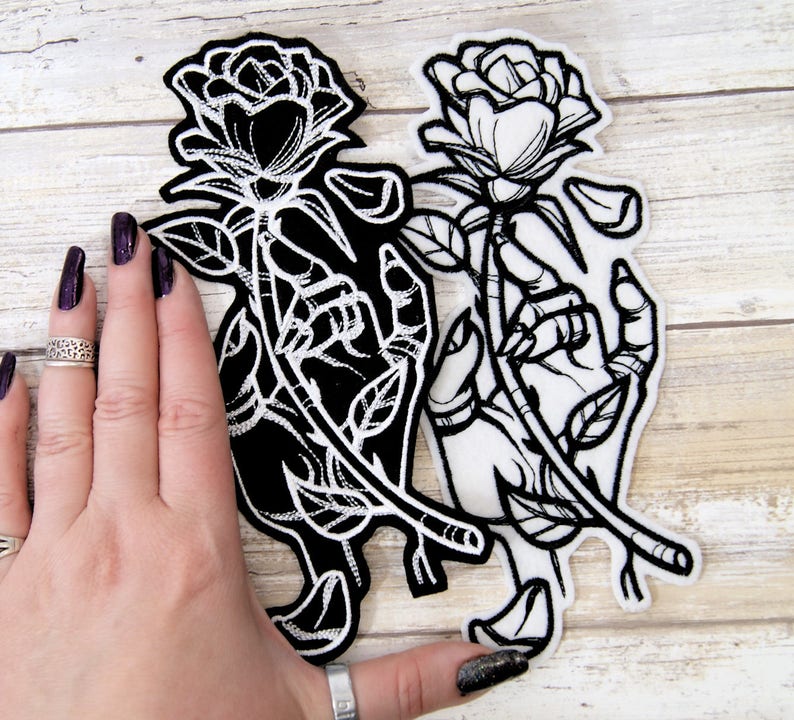 May include: Two black and white embroidered iron-on patches featuring a hand holding a rose. The rose on the left patch is black and the rose on the right patch is white.