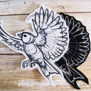 May include: Black and white embroidered design of a bird with its wings spread. The design is on a white fabric background and a black fabric background.