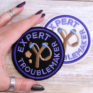 May include: Two embroidered patches with purple stitching. One patch is black with white text that reads "Expert Troublemaker" and a brown slingshot. The other patch is white with purple text that reads "Expert Troublemaker" and a brown slingshot.