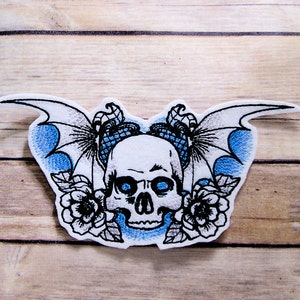 May include: Embroidered iron-on patch featuring a white skull with blue eyes, surrounded by two black wings and two black roses on a blue background.