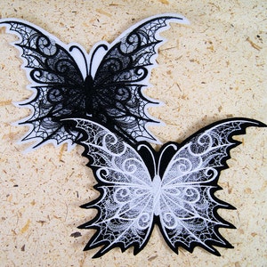 May include: Two black and white butterfly appliques with intricate lace designs. The butterflies are embroidered with a delicate pattern and have a gothic style.