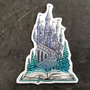 May include: A white embroidered patch with a blue and purple castle on a book. The castle has many towers and a winding path leading up to it. There are green trees on either side of the path.