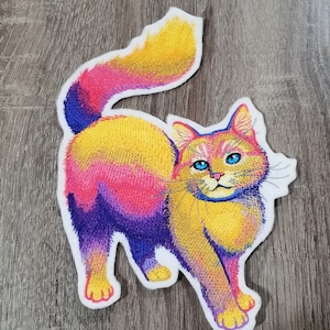 May include: A colorful embroidered patch of a cat with blue eyes. The cat is yellow, pink, and purple.