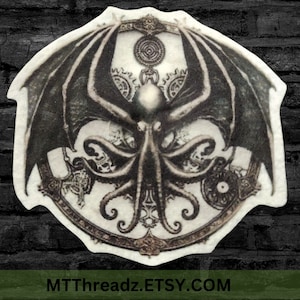May include: A monochrome graphic featuring a winged, tentacled creature within a decorative frame of gears. The image includes the text "MTThreadz.ETSY.COM" at the bottom.