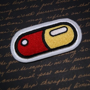 May include: White embroidered patch of a red and yellow pill. The pill is outlined in black.