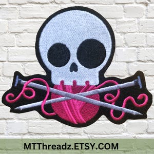 May include: Embroidered iron-on patch featuring a white skull with black eyes and a pink ball of yarn with two crossed knitting needles.