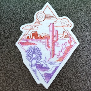 Peut inclure: Écusson brodé représentant un paysage désertique avec un cactus, des montagnes et un crâne de vache. Le motif utilise des fils orange, violets et blancs, encadré dans un losange. Pour vêtements ou accessoires.