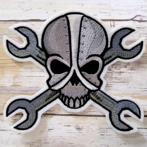 May include: An embroidered patch featuring a grey and white skull wearing a helmet with two crossed spanners behind it.