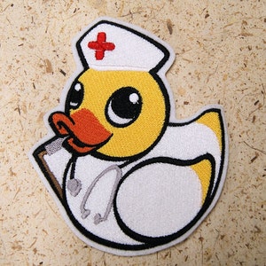 May include: A yellow rubber duck wearing a white nurse's hat with a red cross. The duck is holding a stethoscope and a clipboard.