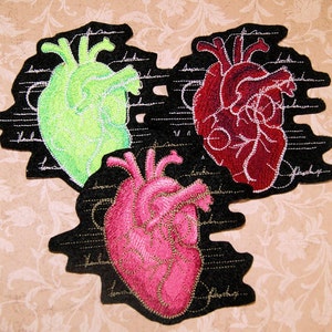 May include: Three embroidered felt patches depicting anatomical hearts. The hearts are green, red, and pink, and are set against a black background with white anatomical markings.