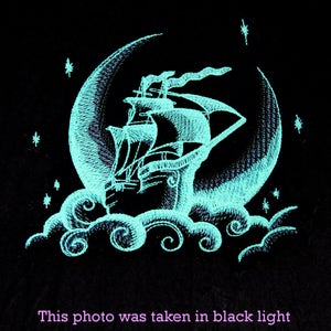 May include: An embroidered design of a sailing ship inside a crescent moon, surrounded by clouds and stars. The design is in a bright teal color against a black background. The text at the bottom reads, "This photo was taken in black light."