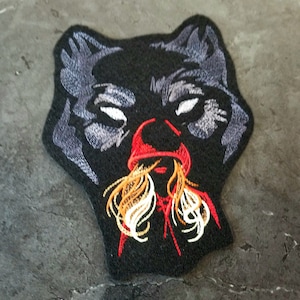 May include: Embroidered patch featuring a black wolf's head with a red hooded figure inside. The wolf's eyes are white and the figure's hair is yellow and white.