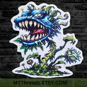 May include: A blue and green cartoon monster plant with a large mouth and sharp teeth. The plant is on a white background with the text "MTTThreadz.ETSY.COM" at the bottom.