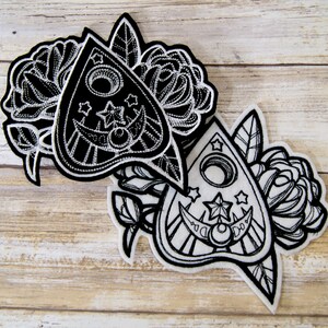 May include: Two black and white embroidered patches featuring a stylized Ouija board with a crescent moon and stars. The patches are surrounded by floral designs.