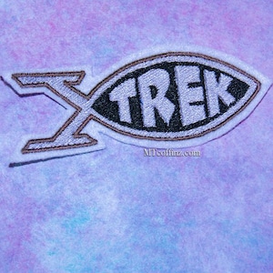 May include: Embroidered patch in the shape of a fish with the word "TREK" in white block letters inside a black oval. The patch has a light purple background and a brown outline. The letter "X" is on the left side of the fish.