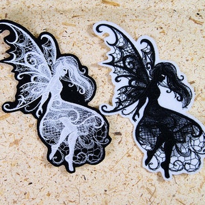 May include: Two embroidered iron-on patches featuring a black and white fairy with wings. The fairy on the left is white with black details and the fairy on the right is black with white details.