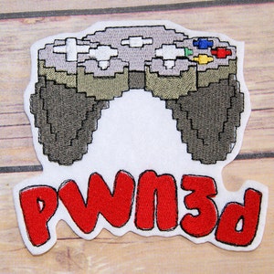 May include: Embroidered patch featuring a pixelated video game controller in shades of gray, green, and white. The patch includes the word "PWN3d" in red, outlined in black. The patch is on a white background.
