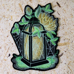 May include: An embroidered patch depicting a black lantern with a glowing yellow light inside. The lantern is surrounded by green leaves and a spider web.
