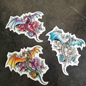 May include: Three colorful dragon tattoos with floral designs. Each dragon is a different color: pink, blue, and green. The dragons are surrounded by flowers and leaves.