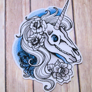 May include: Embroidered patch of a white unicorn skull with a blue and gray gradient background. The skull has a single horn and is adorned with black and white flowers.