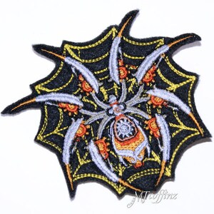 May include: Embroidered iron-on patch featuring a black spider with orange and silver details, sitting in a yellow web on a black background.
