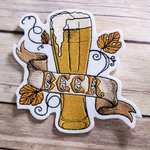 May include: Embroidered patch featuring a beer glass with overflowing foam, surrounded by hops and the word "BEER" on a banner. The patch has a white border and is in shades of gold, brown, and white.