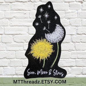 May include: A black, irregularly shaped fabric piece featuring embroidered designs of a bright yellow dandelion, a white dandelion seed head, and white starbursts. The text "Sun, Moon & Stars" is embroidered in white at the bottom.