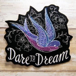 May include: Black felt patch with a purple and blue embroidered swallow flying with white floral details and the text "Dare to Dream" in white embroidery.