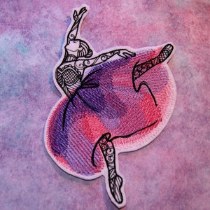 May include: Embroidered iron-on patch of a ballerina in a pink and purple dress with black outlines. The ballerina is in a pose with one leg extended and the other leg bent.