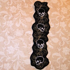 Peut inclure: Garniture en tissu noir avec un bord festonné et un motif répété de crânes blancs et de volutes dorées.